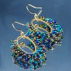 Paire de boucles d'oreilles élégantes de forme oblongue, confectionnées avec des perles multicolores scintillantes et délicates, mêlant des nuances de bleu, vert, violet et doré. Les perles sont minutieusement assemblées sur des arcs métalliques dorés, créant un design harmonieux et sophistiqué. Le crochet d’attache doré ajoute une touche classique et raffinée, parfait pour sublimer une tenue de soirée ou apporter une touche chic au quotidien. Ces bijoux reflètent un artisanat soigné et une attention particulière aux détails.