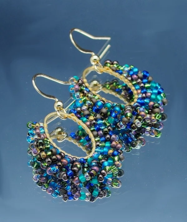 Paire de boucles d'oreilles élégantes de forme oblongue, confectionnées avec des perles multicolores scintillantes et délicates, mêlant des nuances de bleu, vert, violet et doré. Les perles sont minutieusement assemblées sur des arcs métalliques dorés, créant un design harmonieux et sophistiqué. Le crochet d’attache doré ajoute une touche classique et raffinée, parfait pour sublimer une tenue de soirée ou apporter une touche chic au quotidien. Ces bijoux reflètent un artisanat soigné et une attention particulière aux détails.