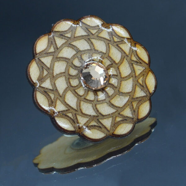 Broche artisanale en bois gravé, décorée d’un motif géométrique inspiré des mandalas et rehaussée d’un cristal facetté aux reflets champagne