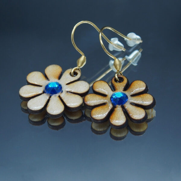 Paire de boucles d’oreilles en forme de marguerites, réalisées en bois finement découpé avec des pétales délicatement définis. Chaque fleur est ornée d’un strass bleu turquoise scintillant en son centre, ajoutant une touche lumineuse et élégante. Montées sur des attaches en acier inoxydable, ces boucles d’oreilles sont légères et confortables à porter.