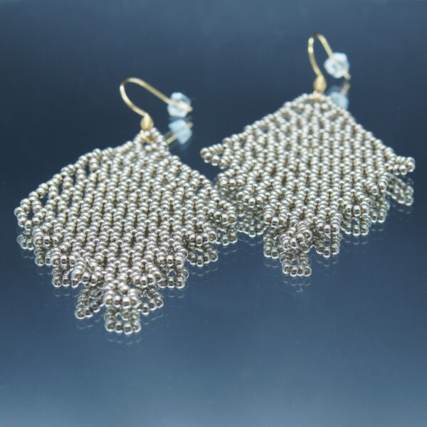 Boucles d'oreilles pendantes en dentelles de perles argentées, formant un motif délicatement tissé. Montées sur des crochets dorés, elles offrent un effet fluide et scintillant, parfait pour une touche d’élégance et de raffinement.