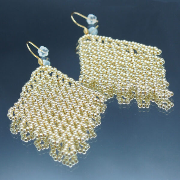 Boucles d'oreilles pendantes en dentelles de perles dorées, formant un motif délicatement tissé. Montées sur des crochets dorés, elles offrent un effet fluide et scintillant, parfait pour une touche d’élégance et de raffinement.